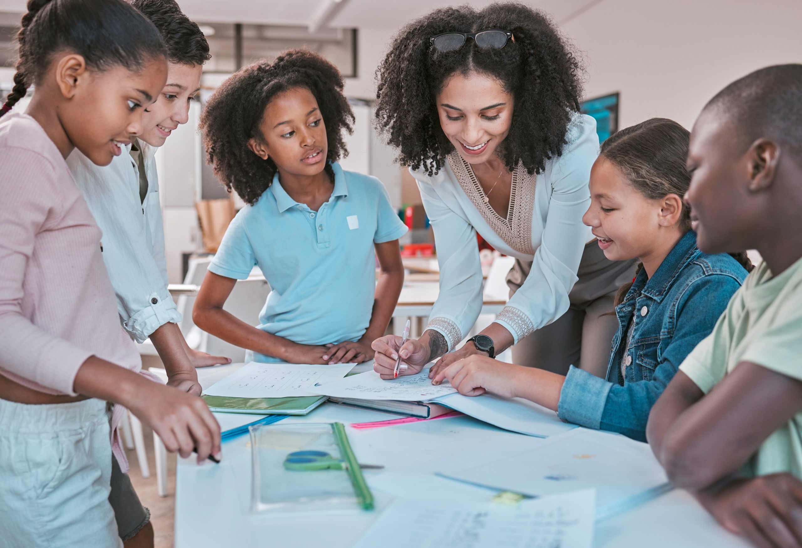 Female teacher in classroom with students, helping learner with homework and writing in book. Childrens education at school, educator reading kids notebook and group learning together for assessment AdobeStock 564943195 scaled